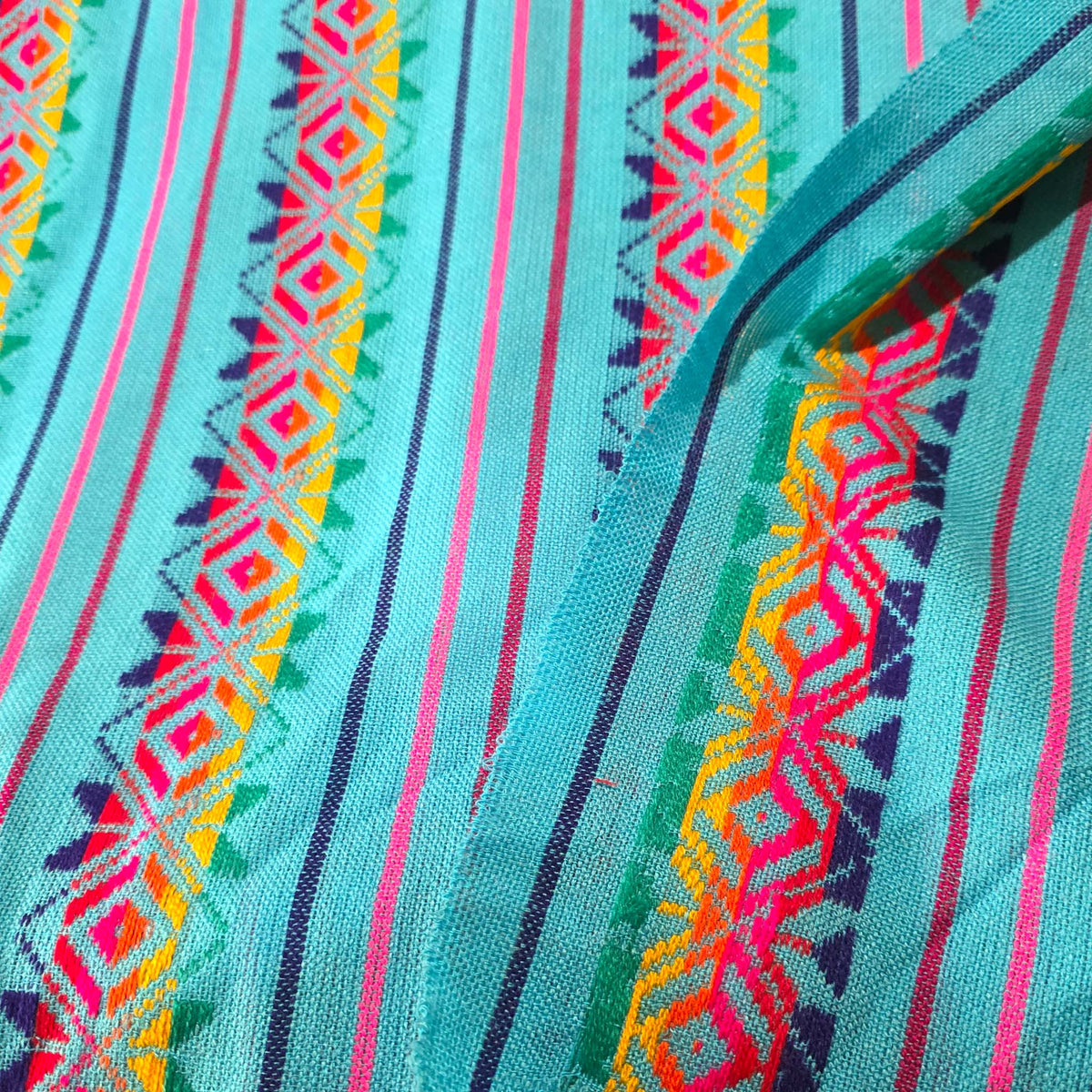 A close-up of a turquoise blue patterned fabric with vertical stripes and colorful geometric embroidery in pink, yellow, green, and purple.