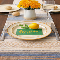 Easter table setting with a plate, lemon, and 'Happy Easter!' card on a striped tablecloth.