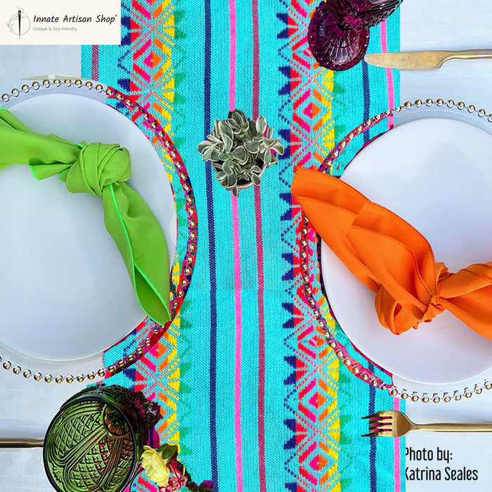 A vibrant turquoise and pink patterned table runner laid under two white plates with colorful napkins, silver chargers, and silver flatware, in a styled flat-lay setting.