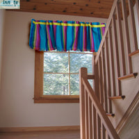 A colorful striped fabric valance with fringe, hanging in front of a window in a rustic room with a wooden staircase and ceiling.