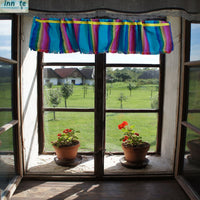 A colorful striped fabric valance with fringe trim hangs above a multi-paned window. The valance features vertical stripes of blue, pink, purple, and green with a yellow stripe down the center.